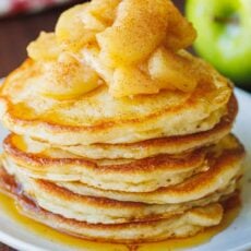 A stack of apple pancakes on a white plate, topped with sauteed cinnamon apples, with green apples in the background.