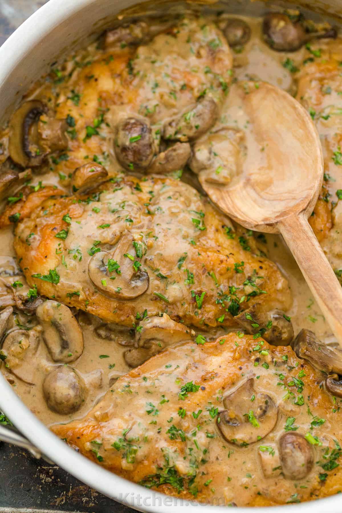 Chicken Marsala in a pan with a wooden spoon topped with mushrooms and parsley.