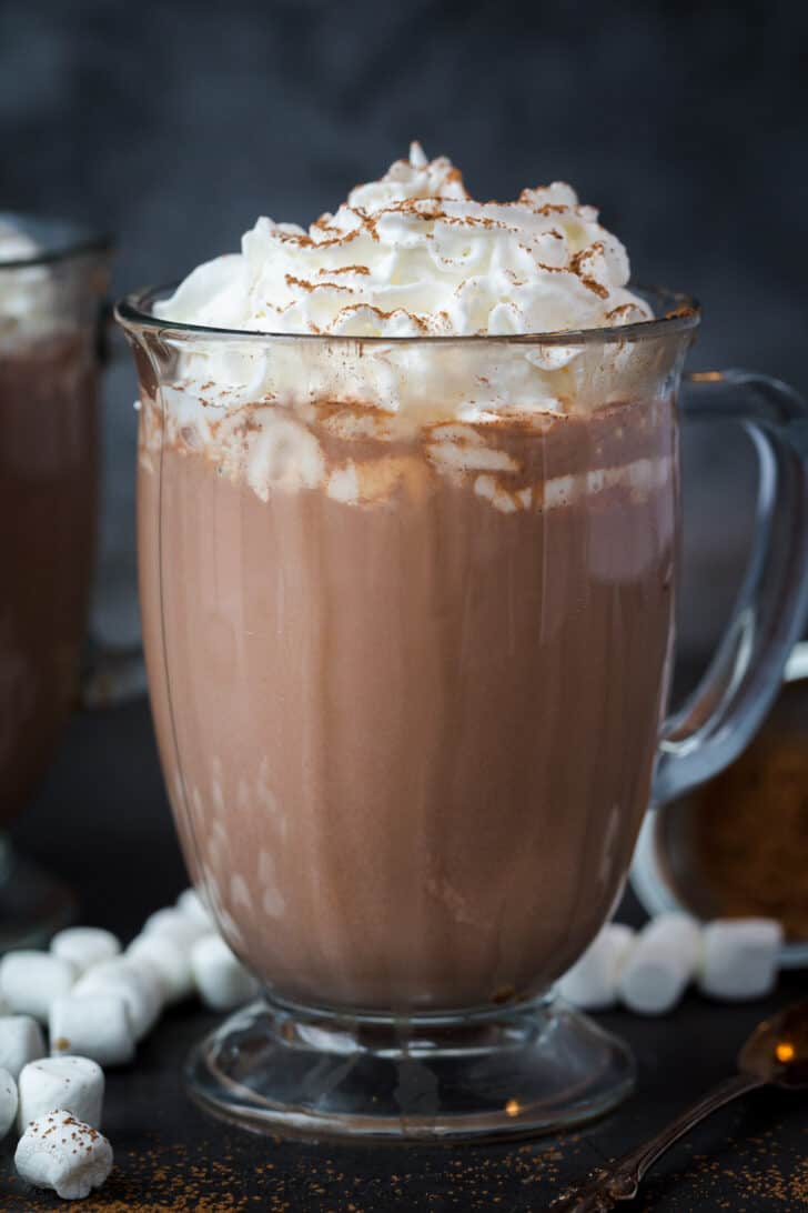 A close up of hot chocolate in a glass mug with whipped cream and cocoa powder.