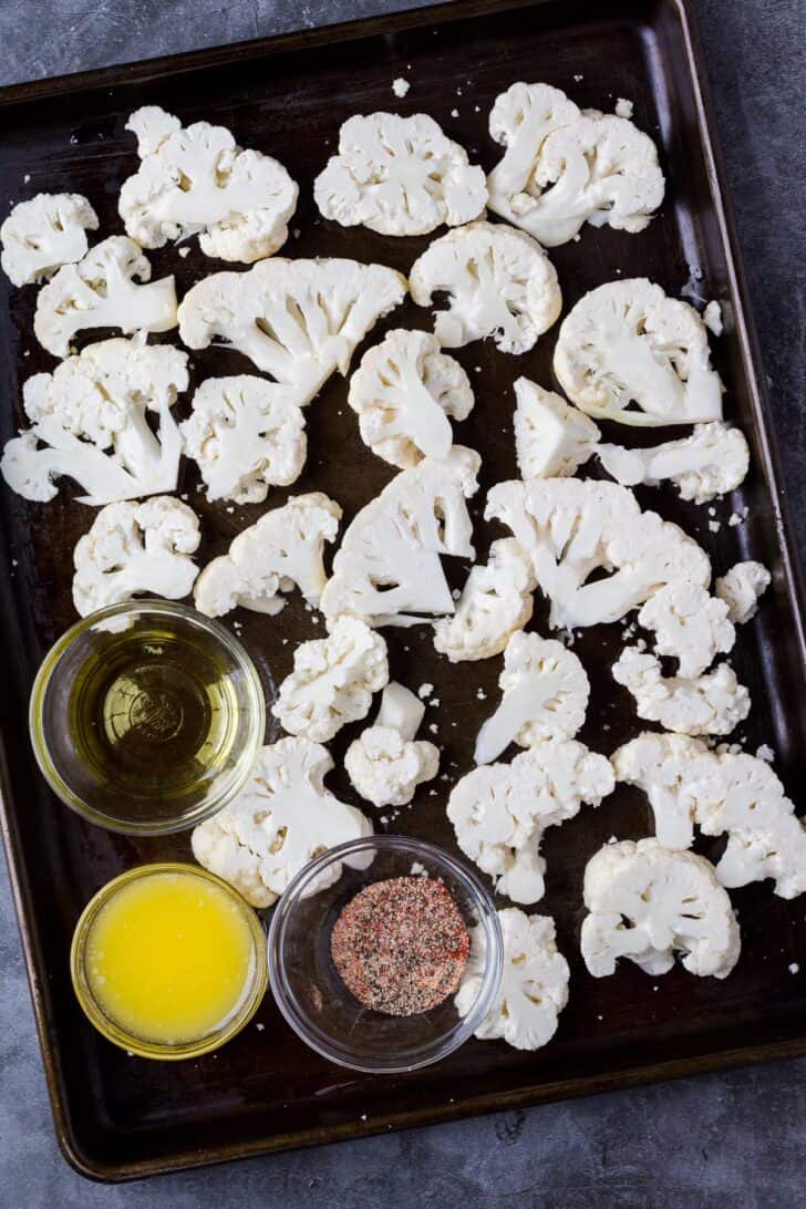 Cauliflower florets on a baking sheet with a glass bowl of oil and another bowl of seasoning. 