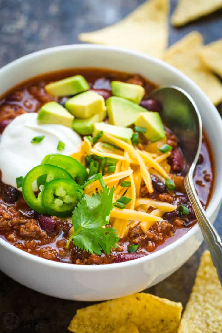 Crockpot chili served in a bowl garnished with toppings and tortilla chips