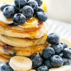 Stack of blueberry pancakes on a plate topped with syrup, blueberries, and sliced bananas.